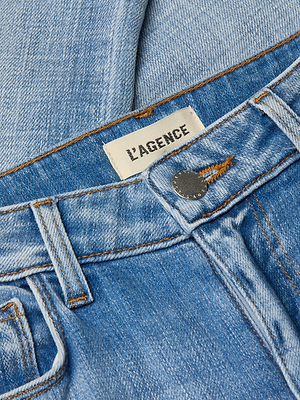 Close-up of the Kendra Cropped Flare Jean in light blue stretch denim, featuring visible stitching, a metal button, front pockets, and an inner waistband label reading L’AGENCE.