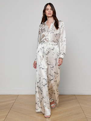 A woman walks indoors on a wooden floor, wearing a silk charmeuse white blouse, Gavin Silk Wide-Leg Pant with a subtle abstract pattern, and open-toed heels.