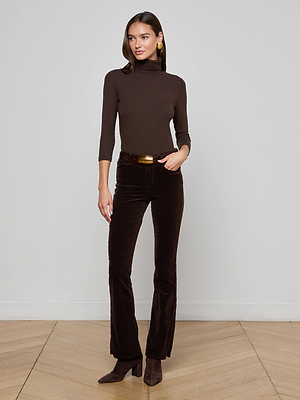 A woman with shoulder-length hair poses indoors against a light gray wall, wearing L'AGENCE's Stevie Velvet Jean in coffee bean brown with a matching turtleneck top and heeled boots on a wooden floor.
