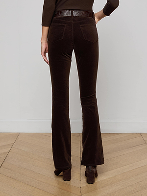 A person faces away, wearing L'AGENCE Stevie Velvet Jean in coffee bean brown, styled with a matching textured belt and heeled shoes. One arm is bent as they stand on a light wooden floor against a plain white wall.