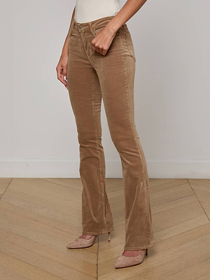 A person wearing the Stevie Velvet Jean in light brown with beige pointed-toe heels stands on a wooden floor against a white wall, showing only their lower body and one arm.