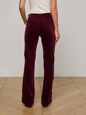 A person stands facing away, wearing L'AGENCE Stevie Velvet Jean—high-waisted, straight-leg maroon stretch velvet pants—with a sleeveless white top. The setting features light wood floors and a plain white wall background.