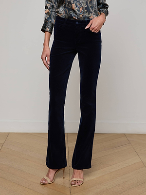 A person wears the L'AGENCE Stevie Velvet Jean in dark navy, paired with a patterned long-sleeve blouse and nude open-toe heels, standing on a wooden floor against a plain light-colored wall.