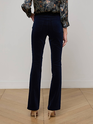 A person stands on a wooden floor, shown from the back, wearing L'AGENCE Stevie Velvet Jeans in dark blue, a patterned long-sleeve blouse, and beige heels.