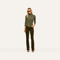 A woman wearing sunglasses, an olive green turtleneck top, and the Stevie Velvet Jean in dark green with a belt stands against a plain light background.