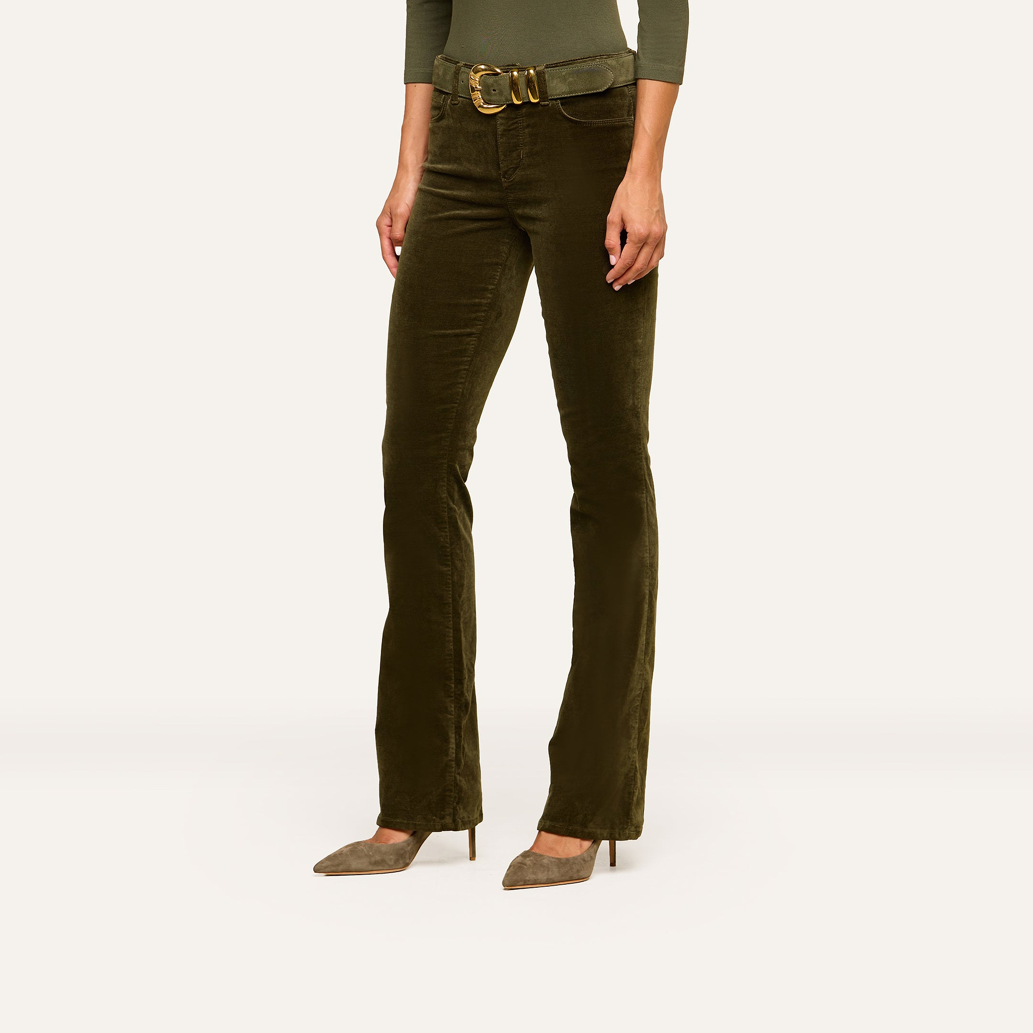 A person wearing Stevie Velvet Jeans in dark green with a matching belt and green pointed-toe heels, cropped from the shoulders down against a plain background.