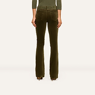 A person stands facing away, wearing the Stevie Velvet Jean—olive-green high rise pants with flared legs and back pockets—paired with heeled shoes. The background is plain and light-colored.