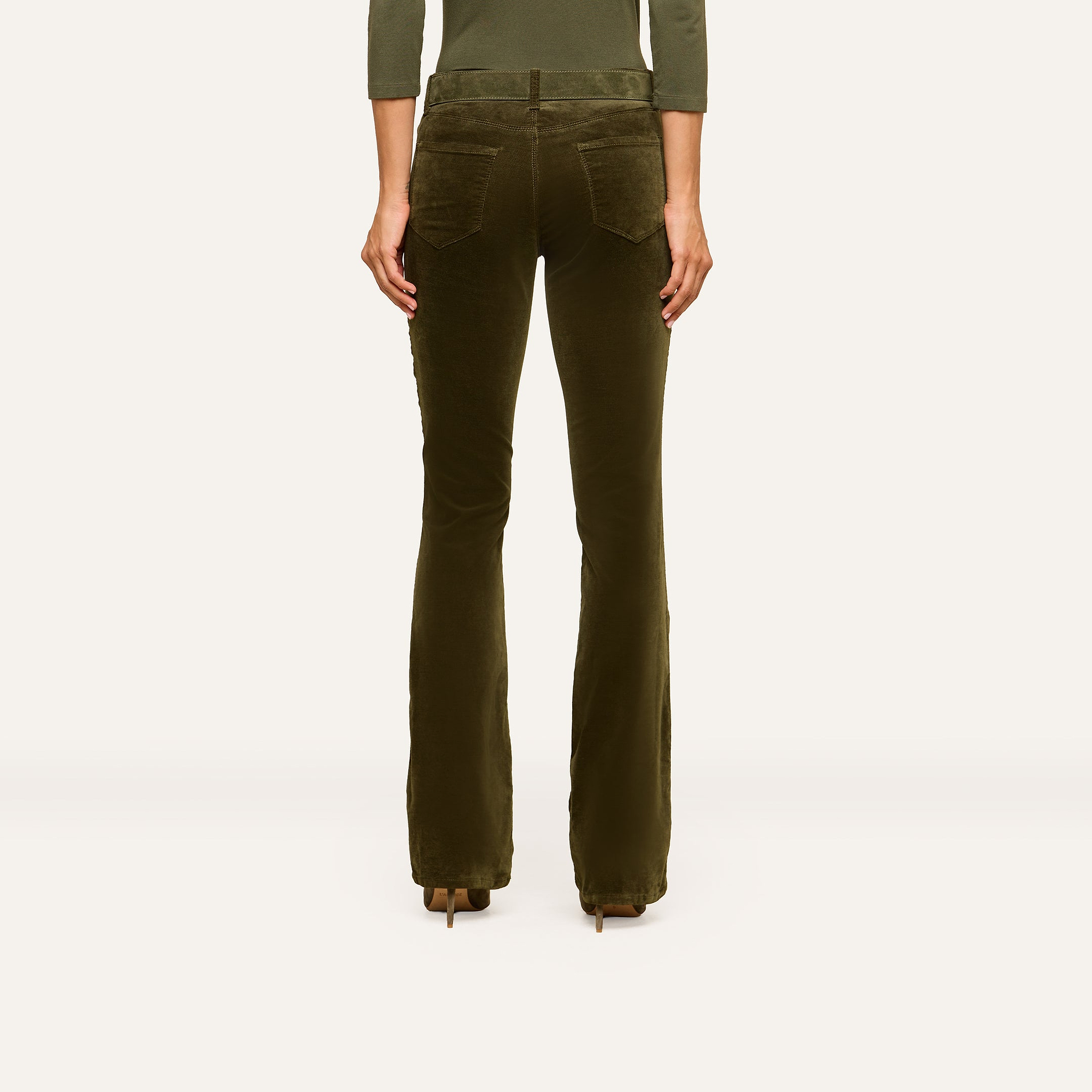 A person stands facing away, wearing the Stevie Velvet Jean—olive-green high rise pants with flared legs and back pockets—paired with heeled shoes. The background is plain and light-colored.
