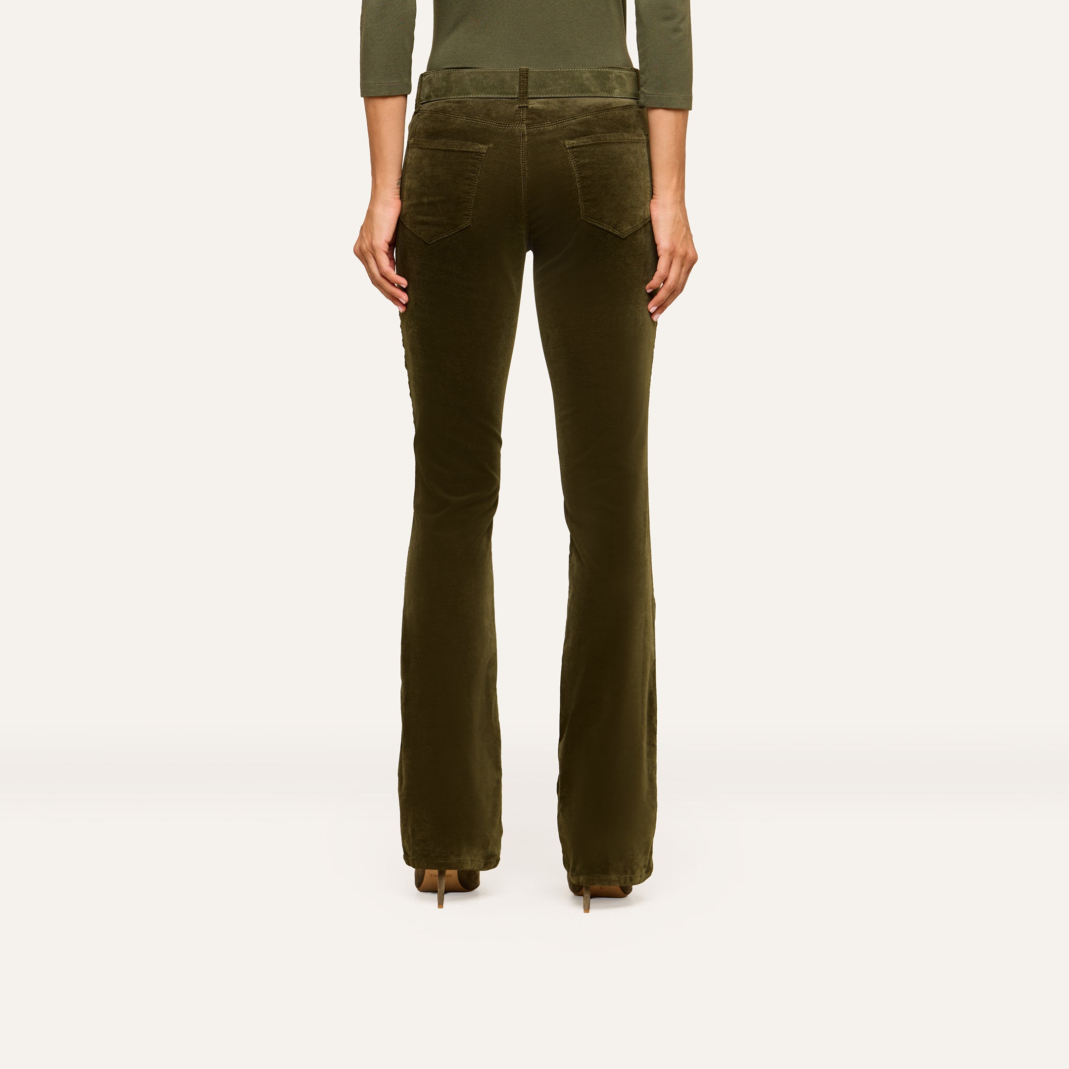 A person stands facing away, wearing the Stevie Velvet Jean—olive-green high rise pants with flared legs and back pockets—paired with heeled shoes. The background is plain and light-colored.
