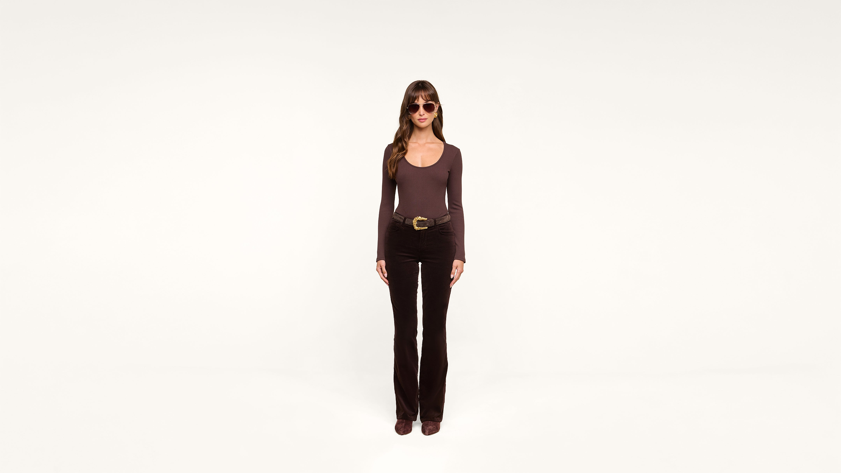 A woman with long brown hair, wearing sunglasses, a brown long-sleeve shirt, black pants, and brown shoes, stands facing forward against a plain white background.