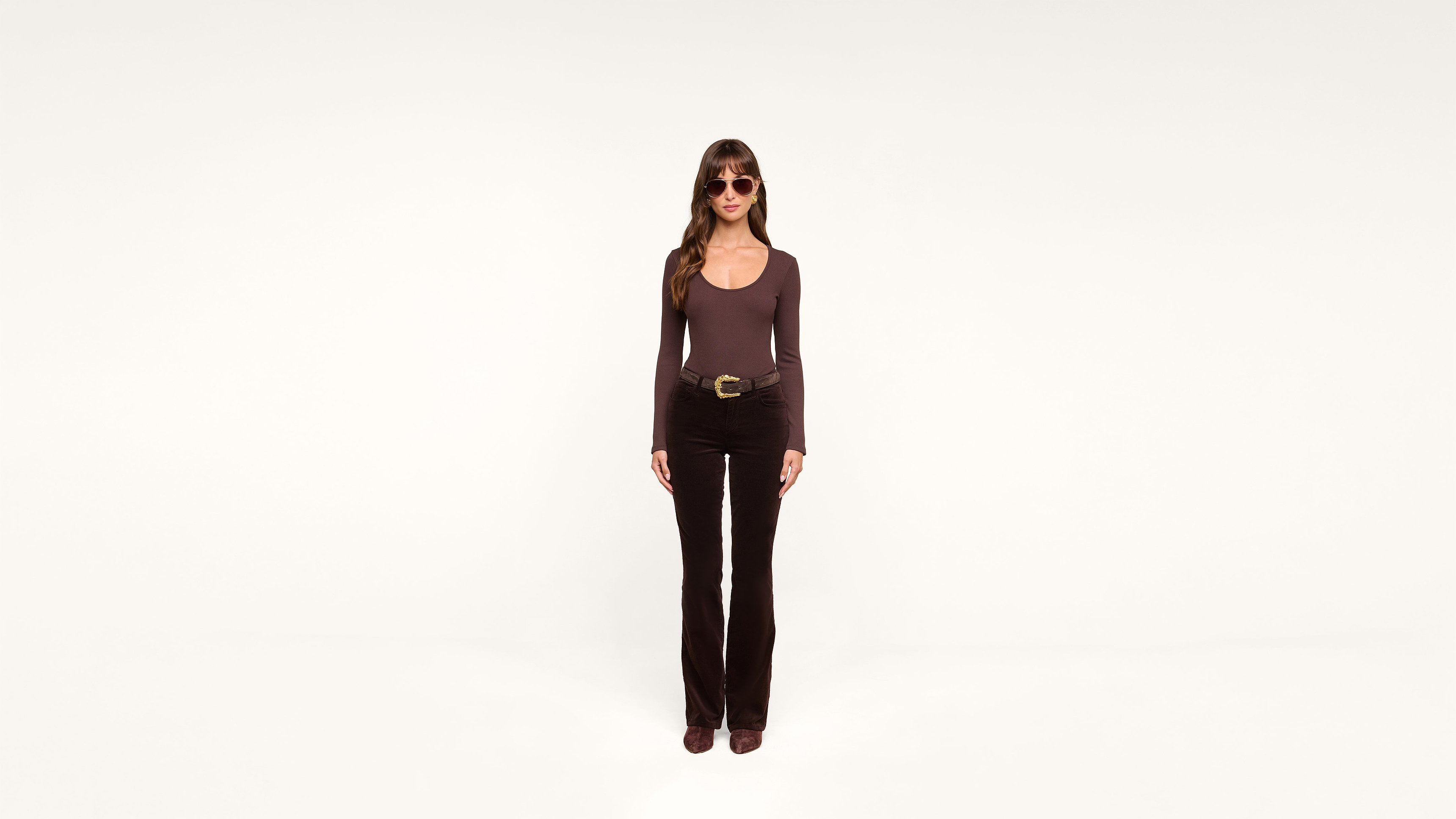 A woman with long brown hair, wearing sunglasses, a brown long-sleeve shirt, black pants, and brown shoes, stands facing forward against a plain white background.
