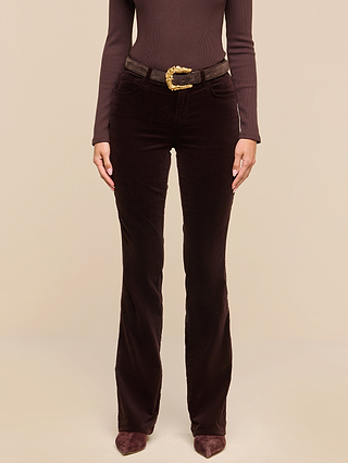 A person wearing a dark java brown long-sleeve top, Stevie Velvet Jean in a slim straight leg fit with stretch fabric and a gold-buckled belt, and brown pointed shoes stands against a beige background.