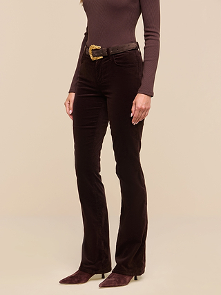 A woman wears the Stevie Velvet Jean in high-waisted, slim straight leg dark java brown stretch velvet, paired with a matching long-sleeve top, a gold-buckled belt, and pointed-toe boots. Only her body from the shoulders down is shown against a beige background.