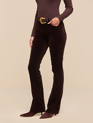 A person wearing a dark brown turtleneck, the Stevie Velvet Jean in dark java brown with a slim straight leg fit, brown pointed-toe boots, and a black belt with a large gold buckle, standing against a plain beige background.