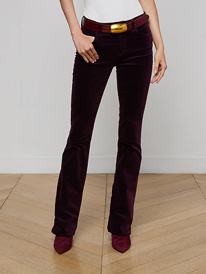 A person stands on a wooden floor against a plain white wall, wearing L'AGENCE Stevie Velvet Jean in dark purple, a white top, a red belt with a gold buckle, and matching pointed shoes.