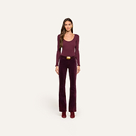 A woman stands facing forward against a plain off-white background, wearing a long-sleeve burgundy top, heeled boots, and high-rise Stevie Velvet Jeans with a belt.
