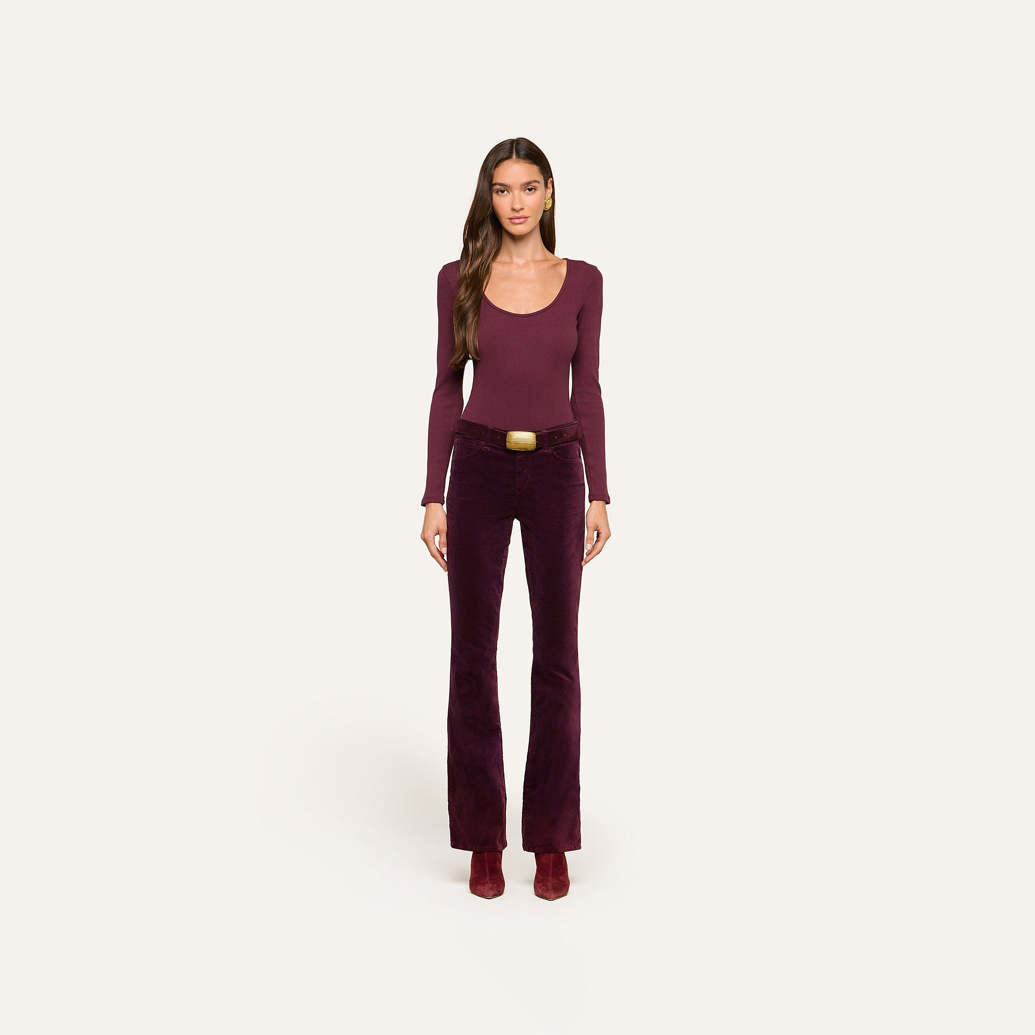 A woman stands facing forward against a plain off-white background, wearing a long-sleeve burgundy top, heeled boots, and high-rise Stevie Velvet Jeans with a belt.