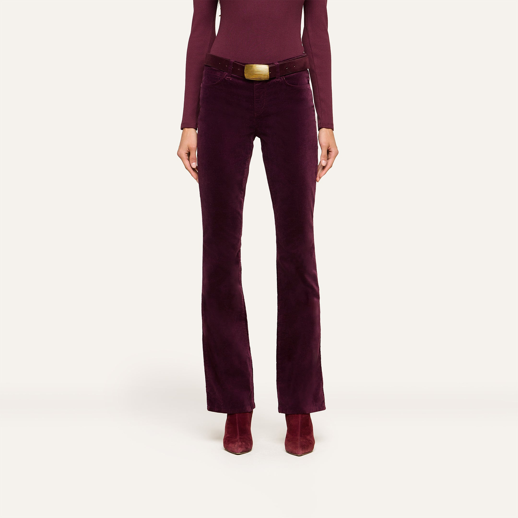 A person wearing a long-sleeved burgundy top, the Stevie Velvet Jean with a matching wide belt featuring a gold buckle, and burgundy heeled boots, standing against a plain light background.