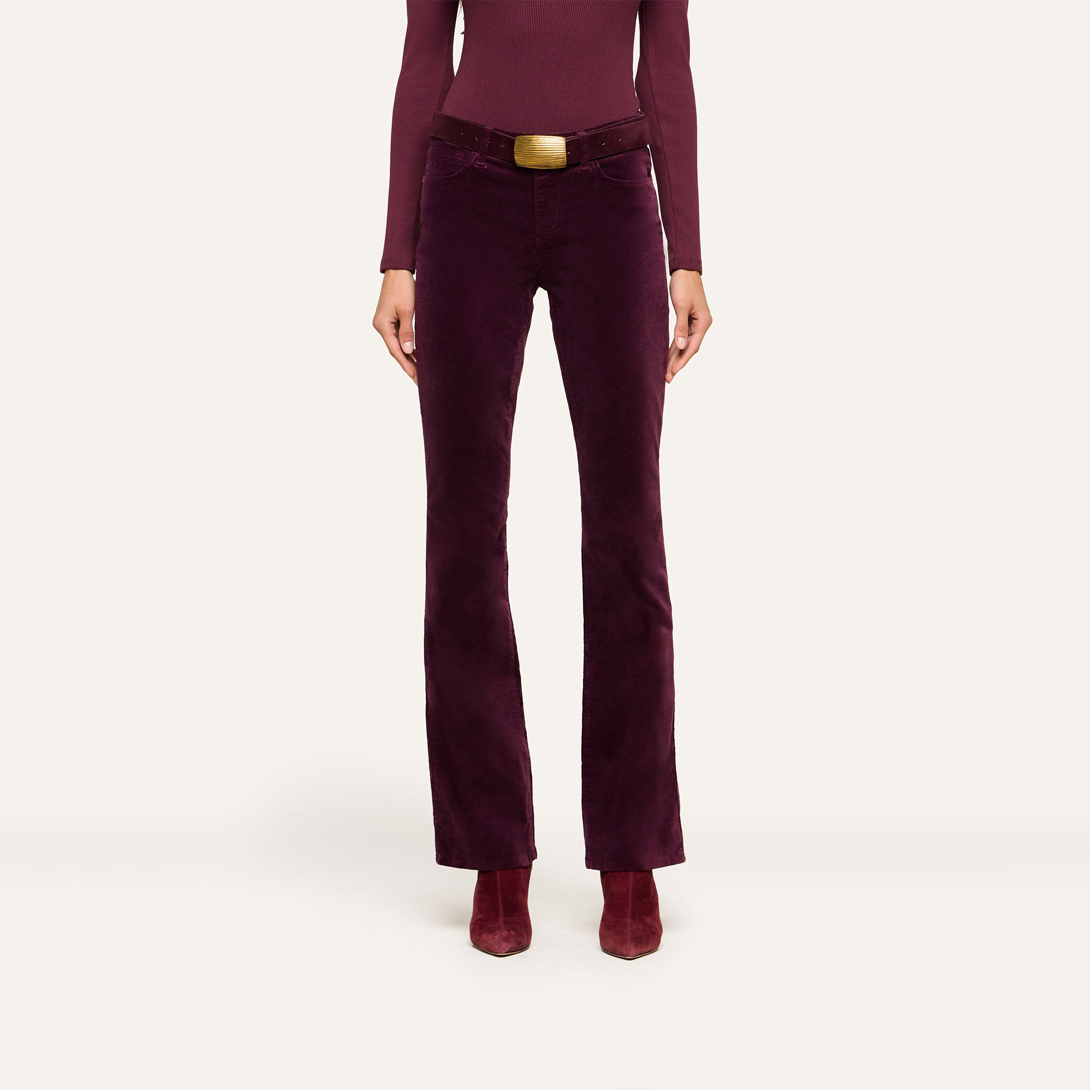 A person wearing a long-sleeved burgundy top, the Stevie Velvet Jean with a matching wide belt featuring a gold buckle, and burgundy heeled boots, standing against a plain light background.
