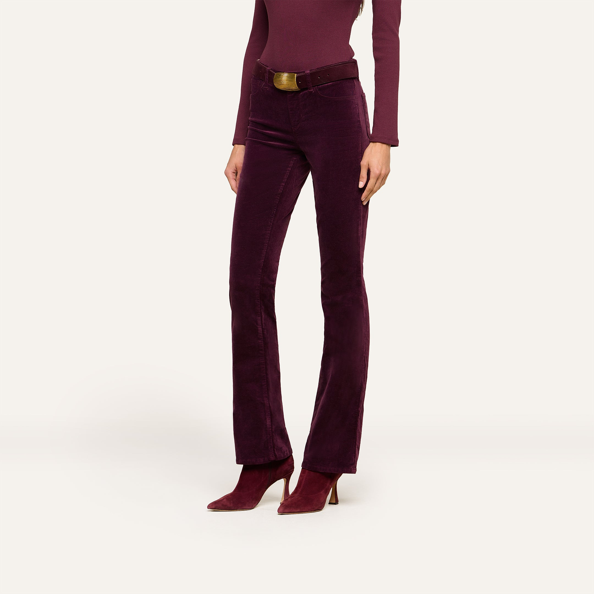 A person wearing a long-sleeve burgundy top, the Stevie Velvet Jean in high-rise dark purple stretch velvet with a belt, and matching pointed-toe heeled boots stands against a plain background.