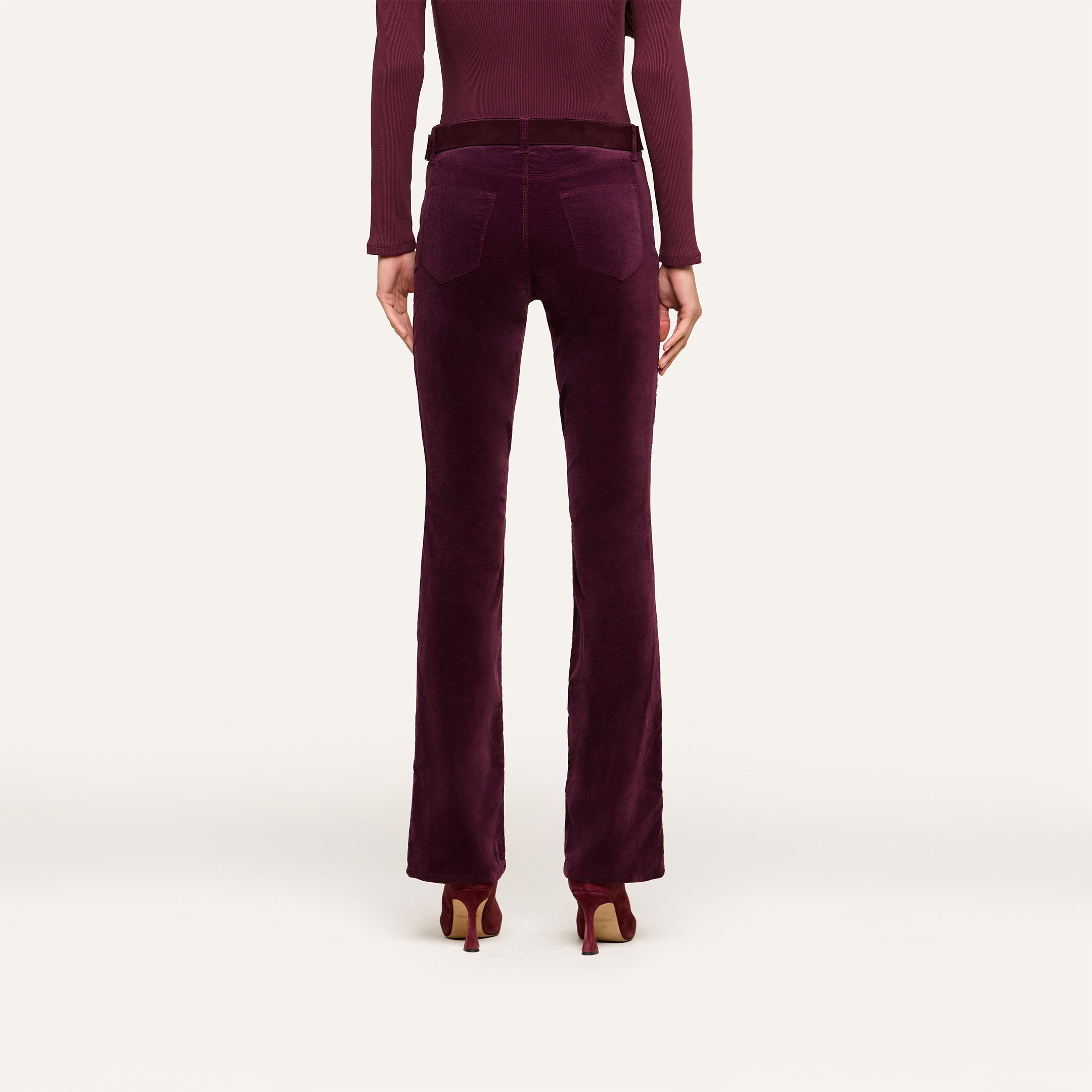 A person wearing Stevie Velvet Jeans in dark burgundy stretch velvet with matching heels, seen from the back against a plain light background; their long-sleeved top is also burgundy.