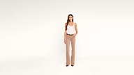 A woman in a white tank top, a brown belt with a gold buckle, and tan trousers stands against a white background.