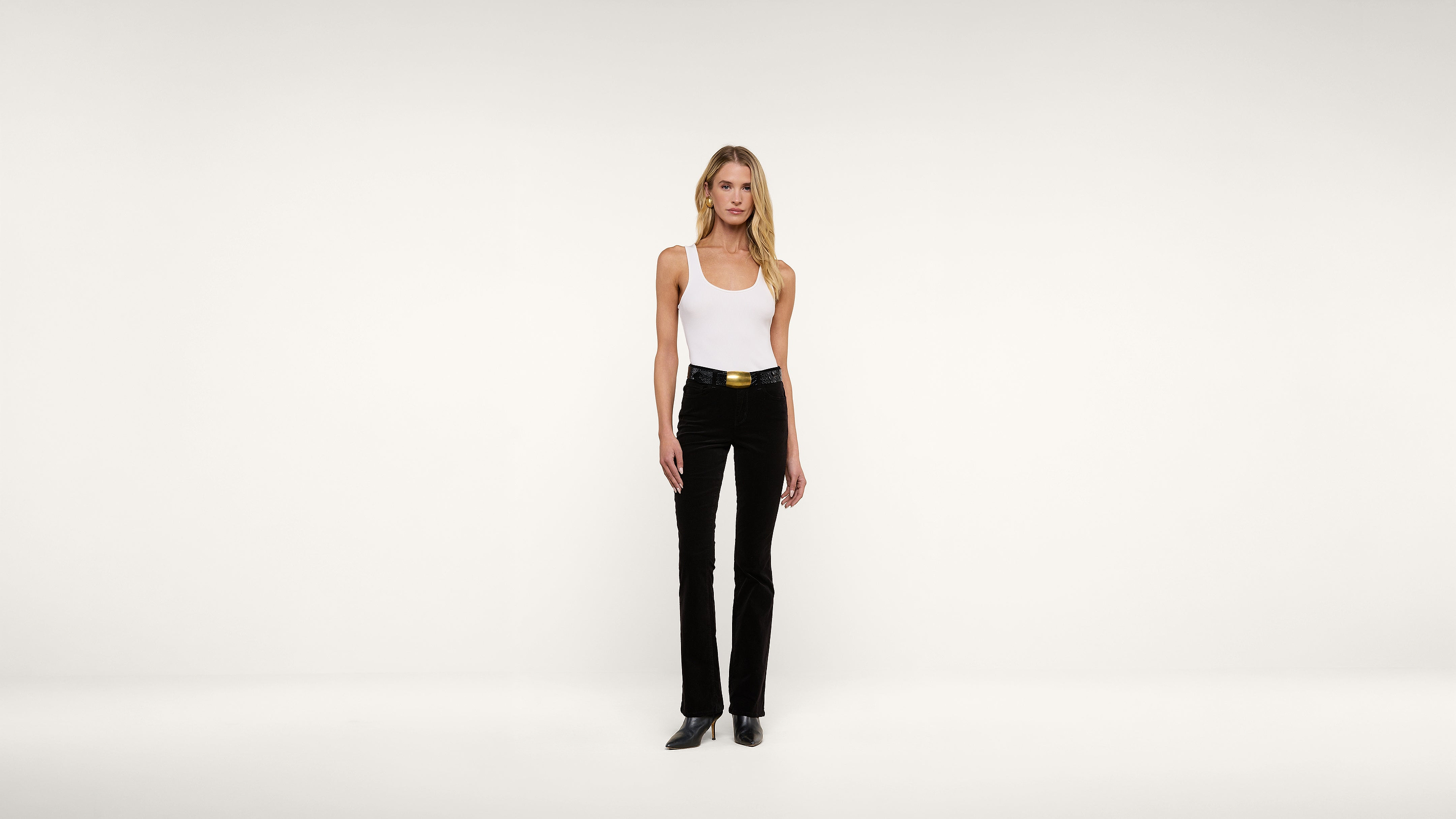 A woman stands against a plain white background, wearing a white tank top, black pants with a belt, and black shoes.