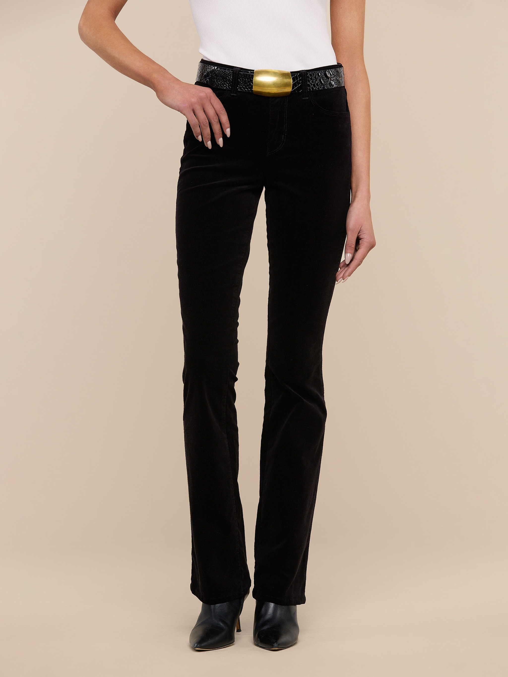 A person wearing a white sleeveless top, the Stevie Velvet Jean in black, black shoes, and a large gold belt buckle stands against a plain white background.