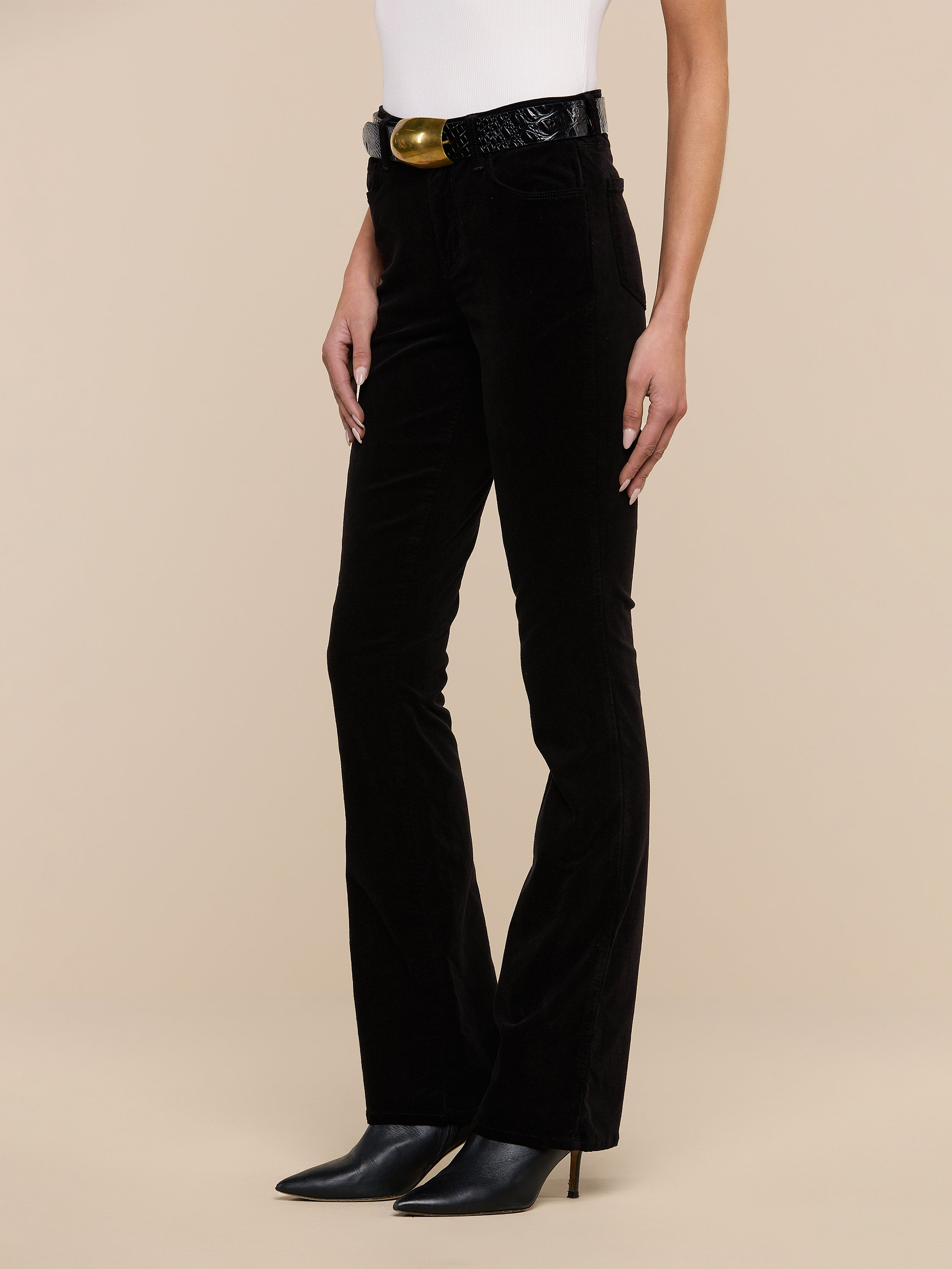 A person wearing the Stevie Velvet Jean in black, paired with a white sleeveless top, a black belt with a gold buckle, and black pointed-toe heeled boots stands against a plain white background.