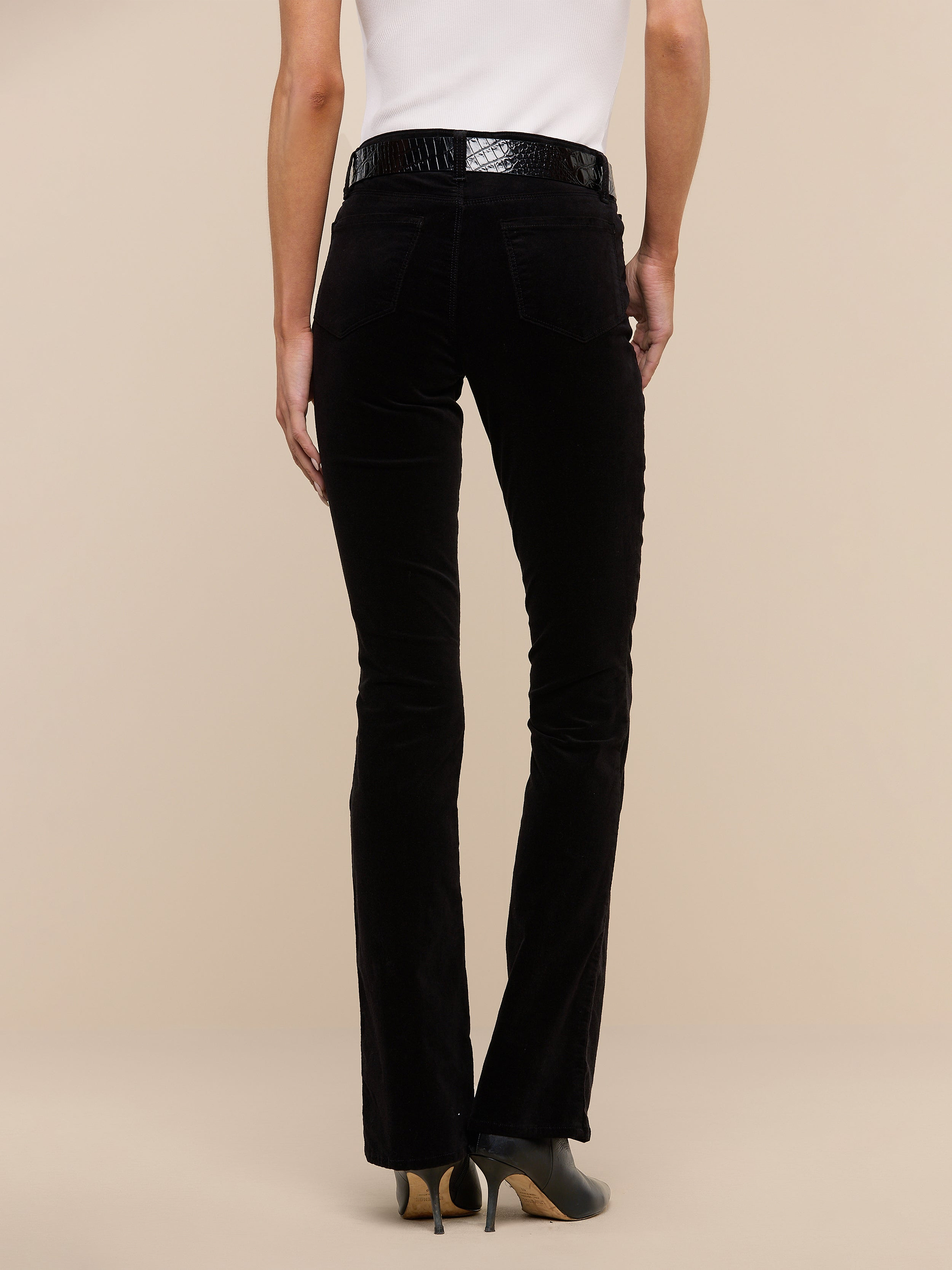 A person wearing black Stevie Velvet Jeans, a white sleeveless top, and black heeled shoes stands facing away against a plain white background.