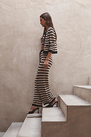 A woman with long brown hair wearing a black and white striped two-piece outfit and black heels walks up concrete stairs against a beige textured wall.