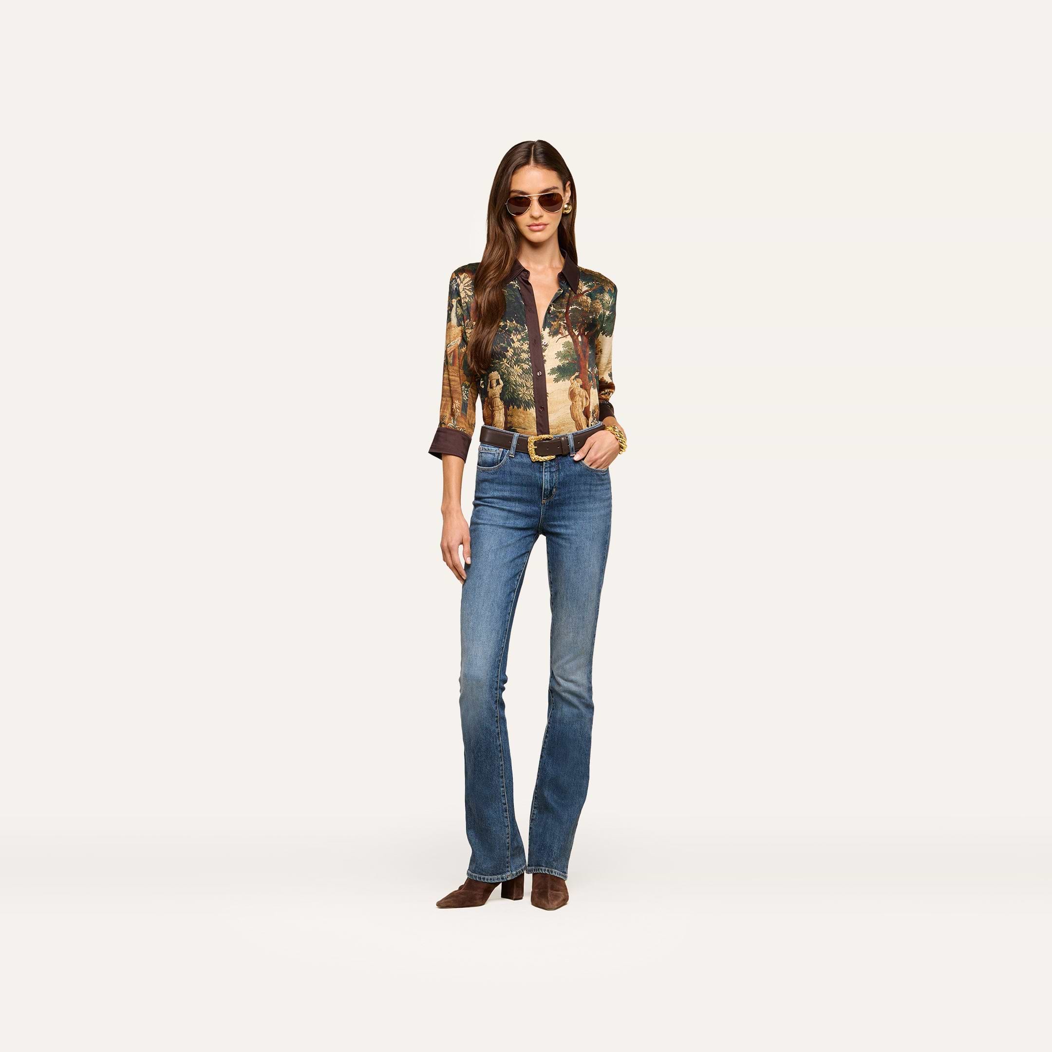 A woman wearing sunglasses, a patterned blouse, the Selma Bootcut Jean crafted from stretch denim, and brown boots stands confidently against a plain light background.