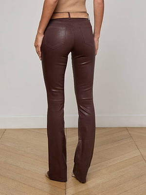 A person stands on a wooden floor, shown from the back, wearing L'AGENCE's Selma Coated Bootcut Jean in chocolate brown. The background is plain and light-colored.