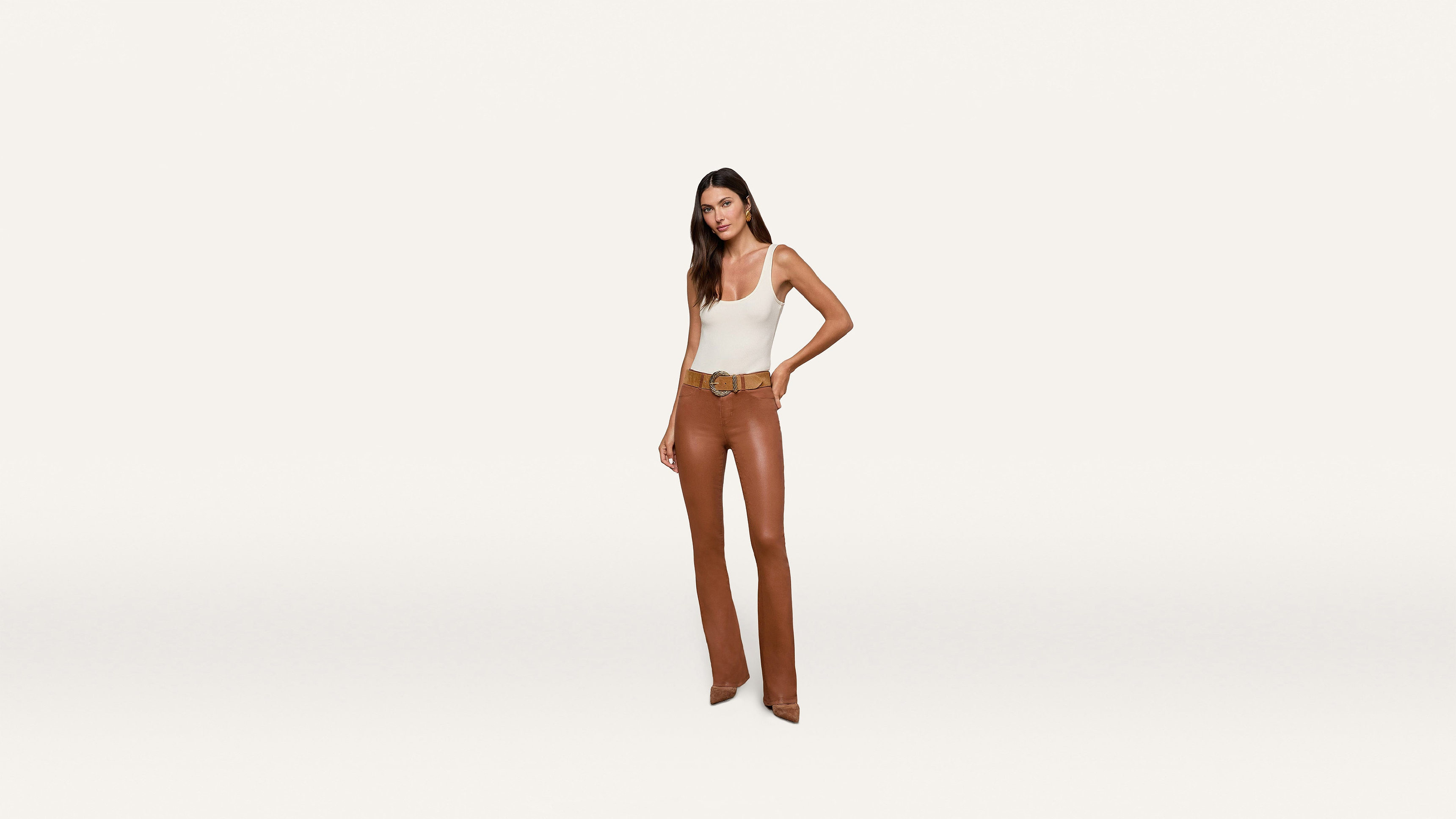 A woman stands against a plain white background, wearing a white sleeveless top paired with brown high-waisted pants, a belt, and brown heels. She has long brown hair and confidently poses with one hand on her hip.