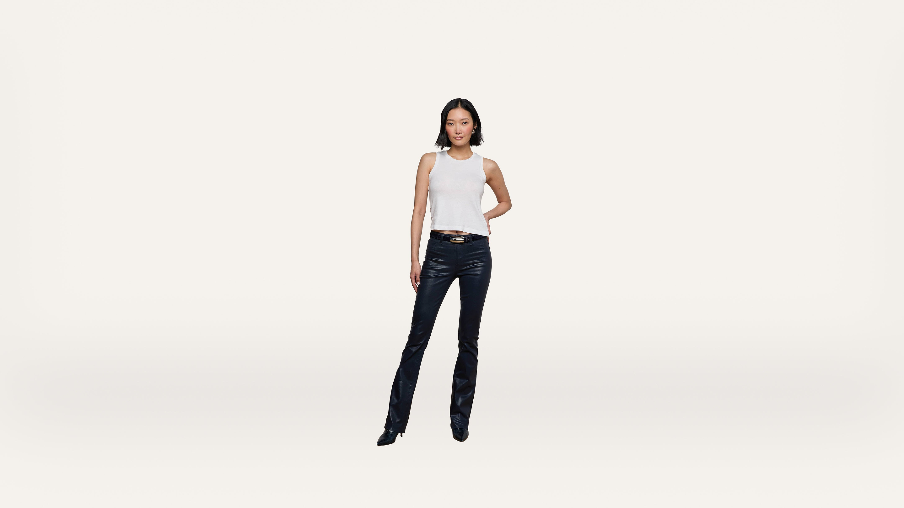 A woman stands confidently against a plain white background, wearing a sleeveless white top, black leather pants, and black shoes.