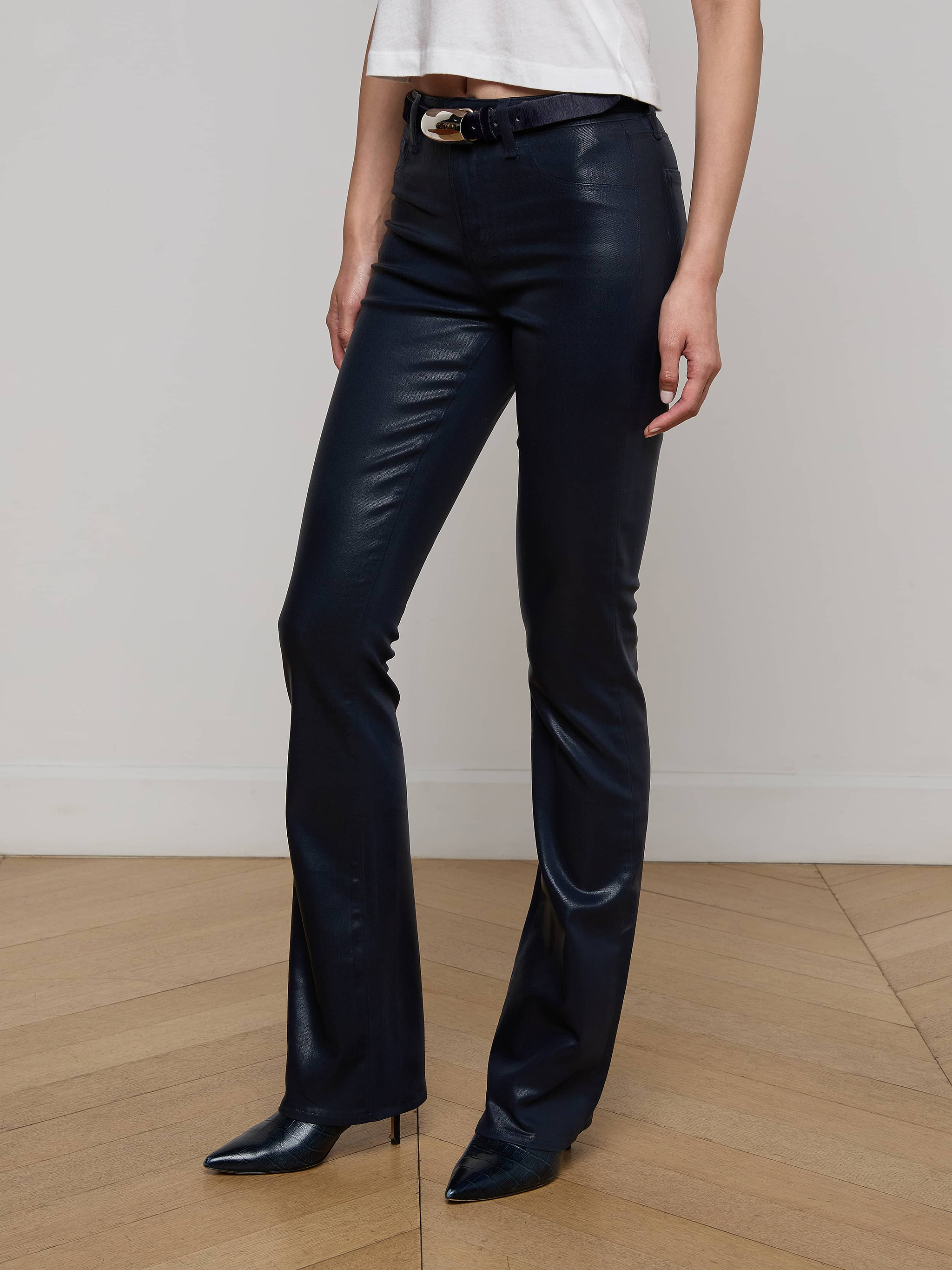 A person wearing the Selma Coated Bootcut Jean, paired with a white top and black pointed-toe shoes, stands against a plain white background. Only the lower half of the body is visible.