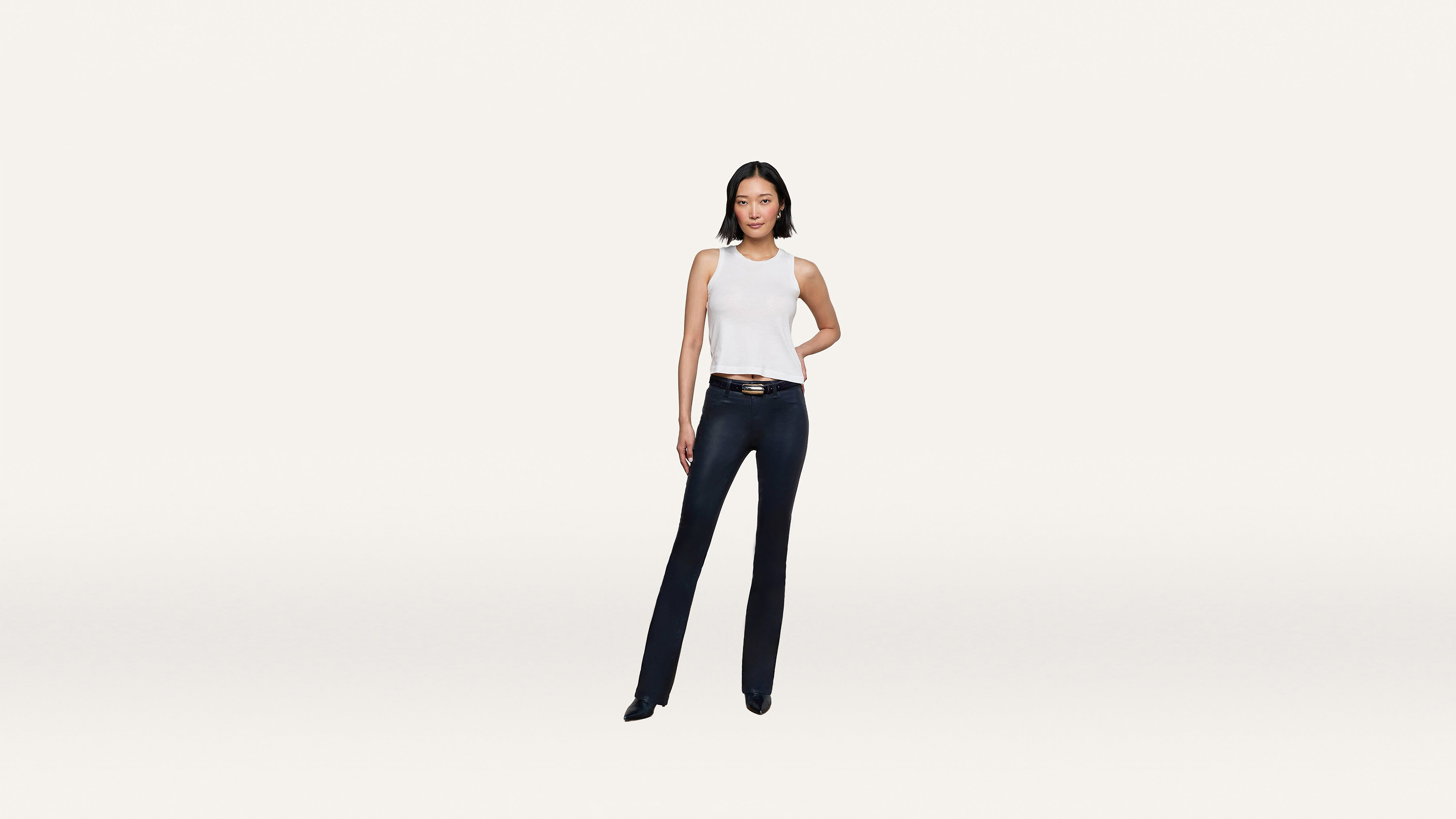 Woman stands facing forward, wearing a crisp white sleeveless top, dark blue jeans, and black shoes, with one hand on her hip against a plain white background.