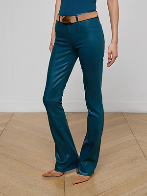 A person stands on a wooden floor wearing L’AGENCE’s Selma Coated Bootcut Jean, a brown belt, and brown pointed-toe shoes. The upper body is partially visible against a plain, light-colored background.