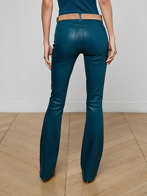 A person stands on a wooden floor, seen from the back, wearing L'AGENCE Selma Coated Bootcut Jeans in teal with a tan belt and dark top. The jeans have a slim fit and are slightly flared at the bottom.