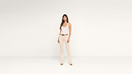 A woman in a white tank top and beige pants with a brown belt and heels stands confidently against a plain white background.