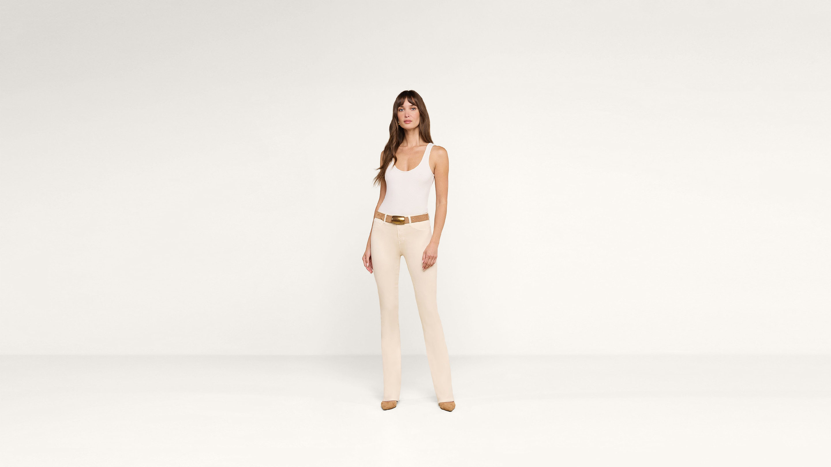 A woman in a white tank top and beige pants with a brown belt and heels stands confidently against a plain white background.