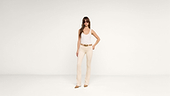 A woman in sunglasses, a white tank top, beige pants, and brown shoes stands against a plain white background with one hand on her hip.