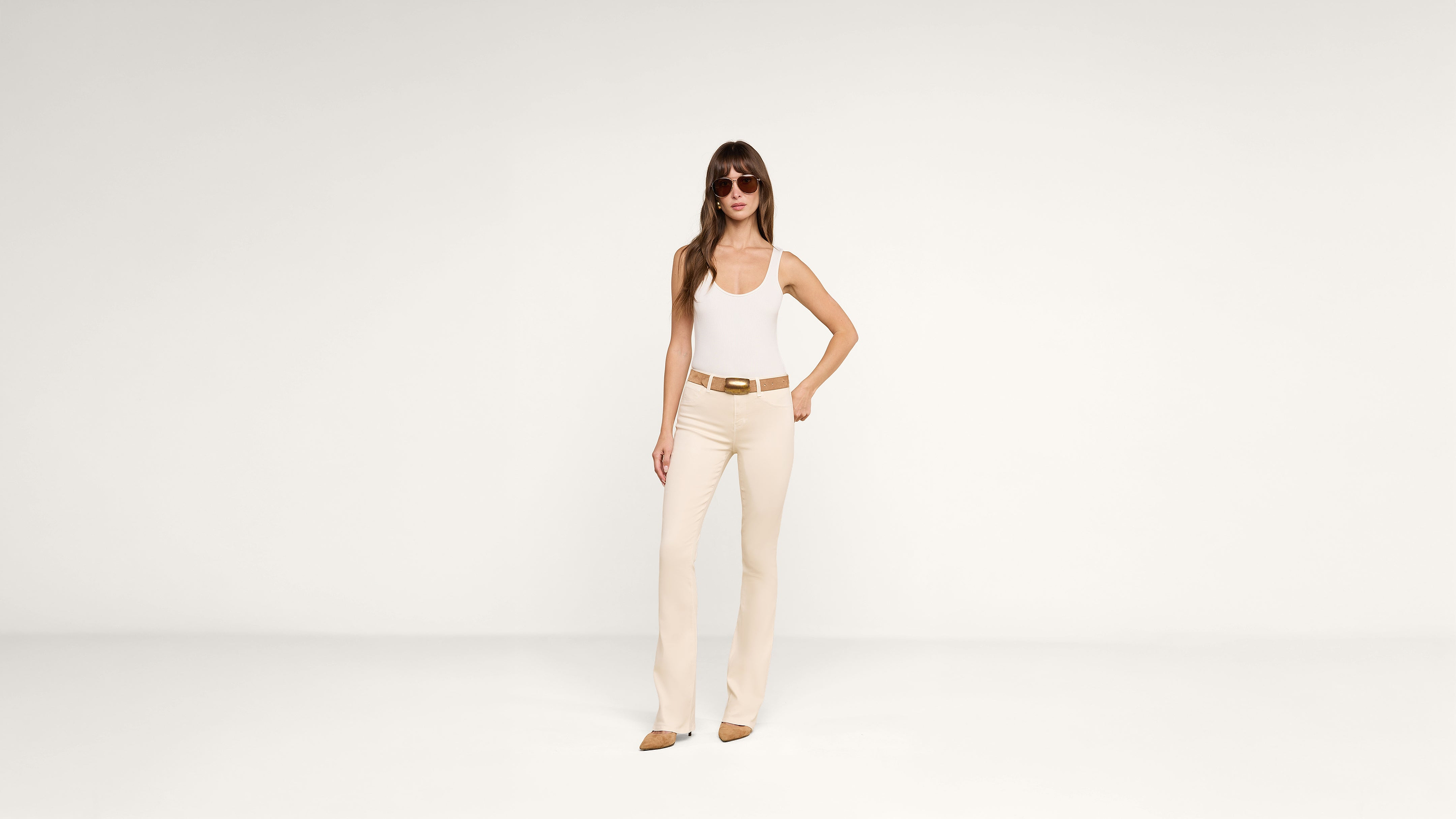 A woman in sunglasses, a white tank top, beige pants, and brown shoes stands against a plain white background with one hand on her hip.