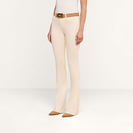 A person wearing the Selma Coated Bootcut Jean in beige, styled with a brown belt, a white top, and brown pointed shoes. They are standing against a plain white background, with their face not visible in the image.