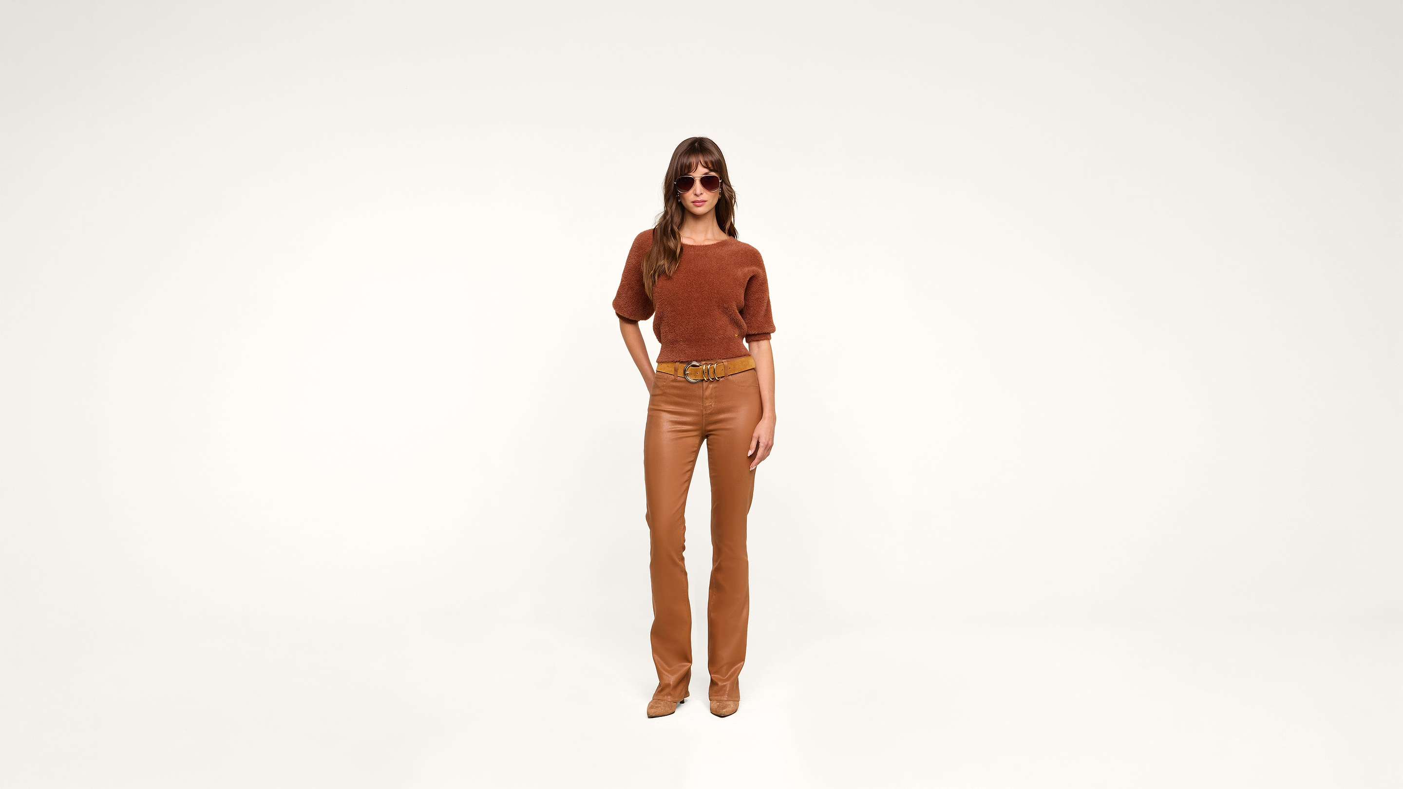 A woman wearing sunglasses, a brown top, tan belt, tan pants, and tan shoes stands against a plain white background, looking confidently at the camera.