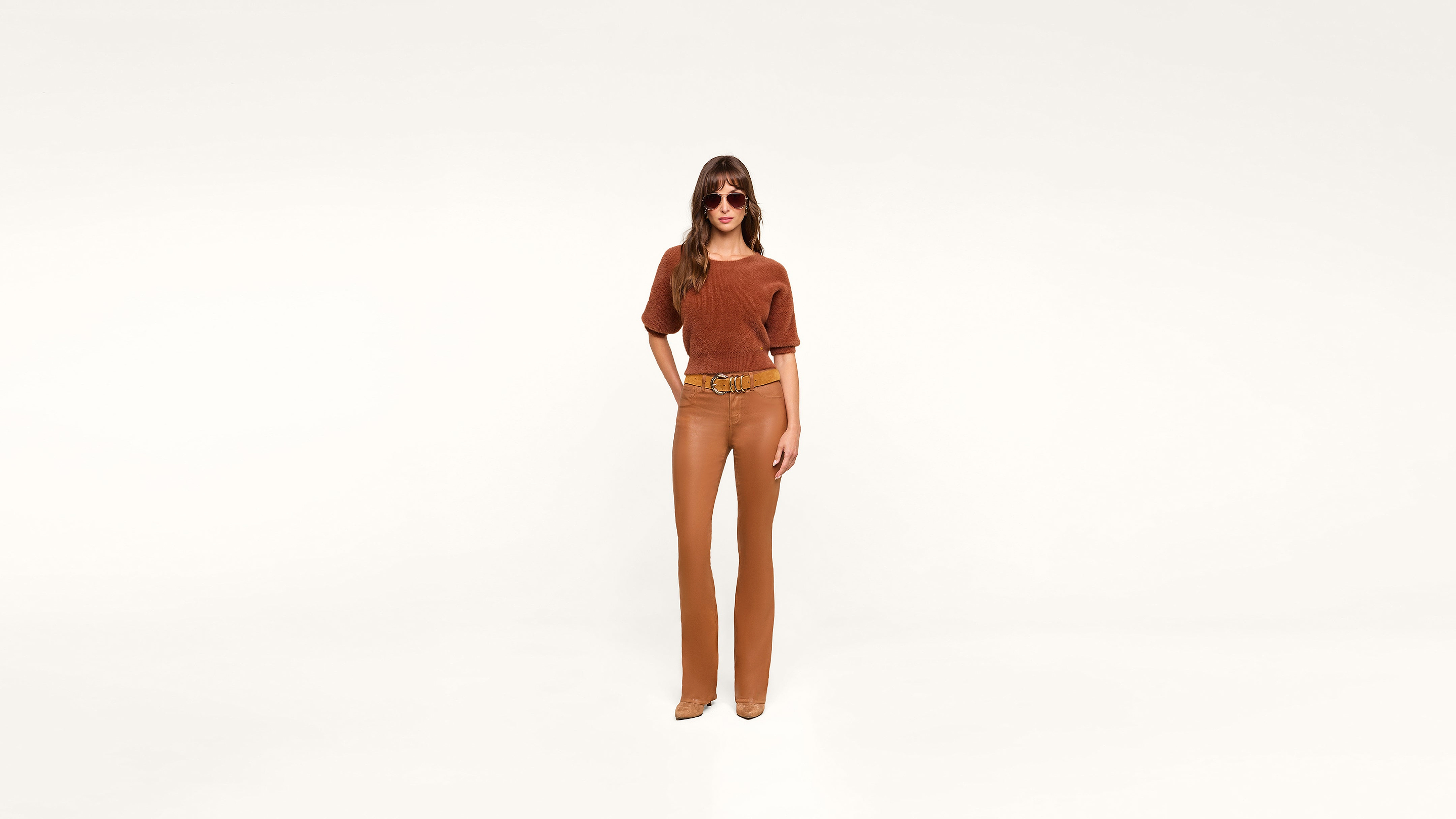 A woman wearing sunglasses, a rust-colored top, tan pants, and tan shoes stands stylishly against a plain white background.