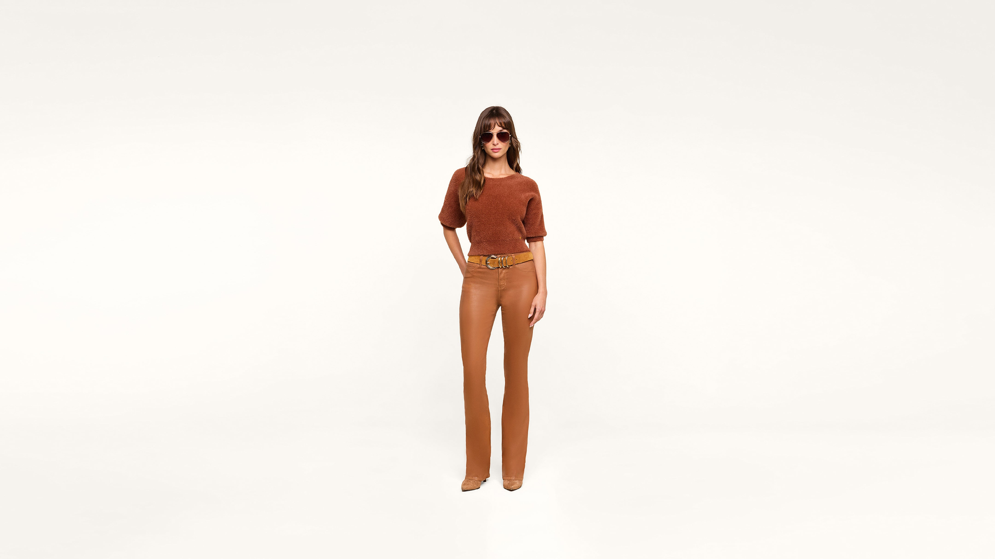 A woman wearing sunglasses, a rust-colored top, tan pants, and tan shoes stands stylishly against a plain white background.