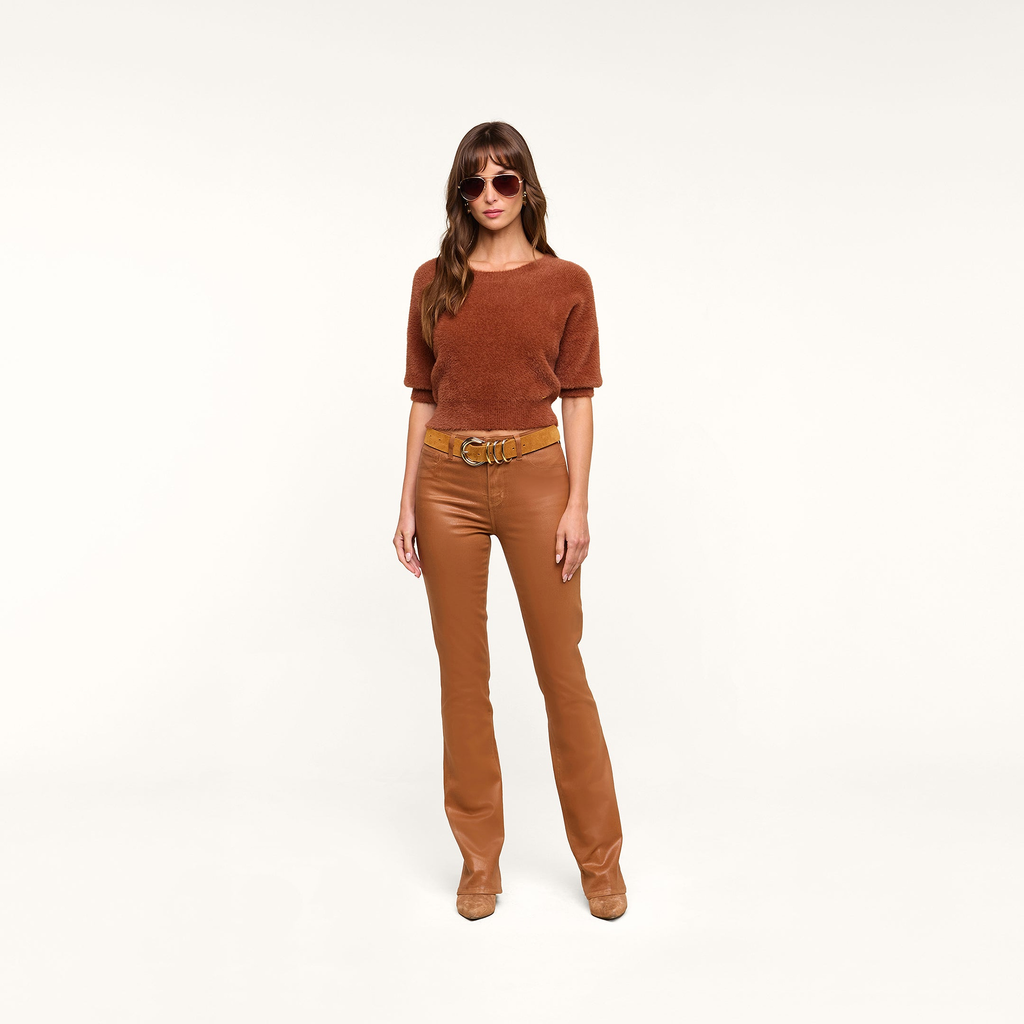 A woman wearing sunglasses, a dark java brown short-sleeve sweater, the Selma Coated Bootcut Jean with a belt, and brown shoes stands against a plain white background.