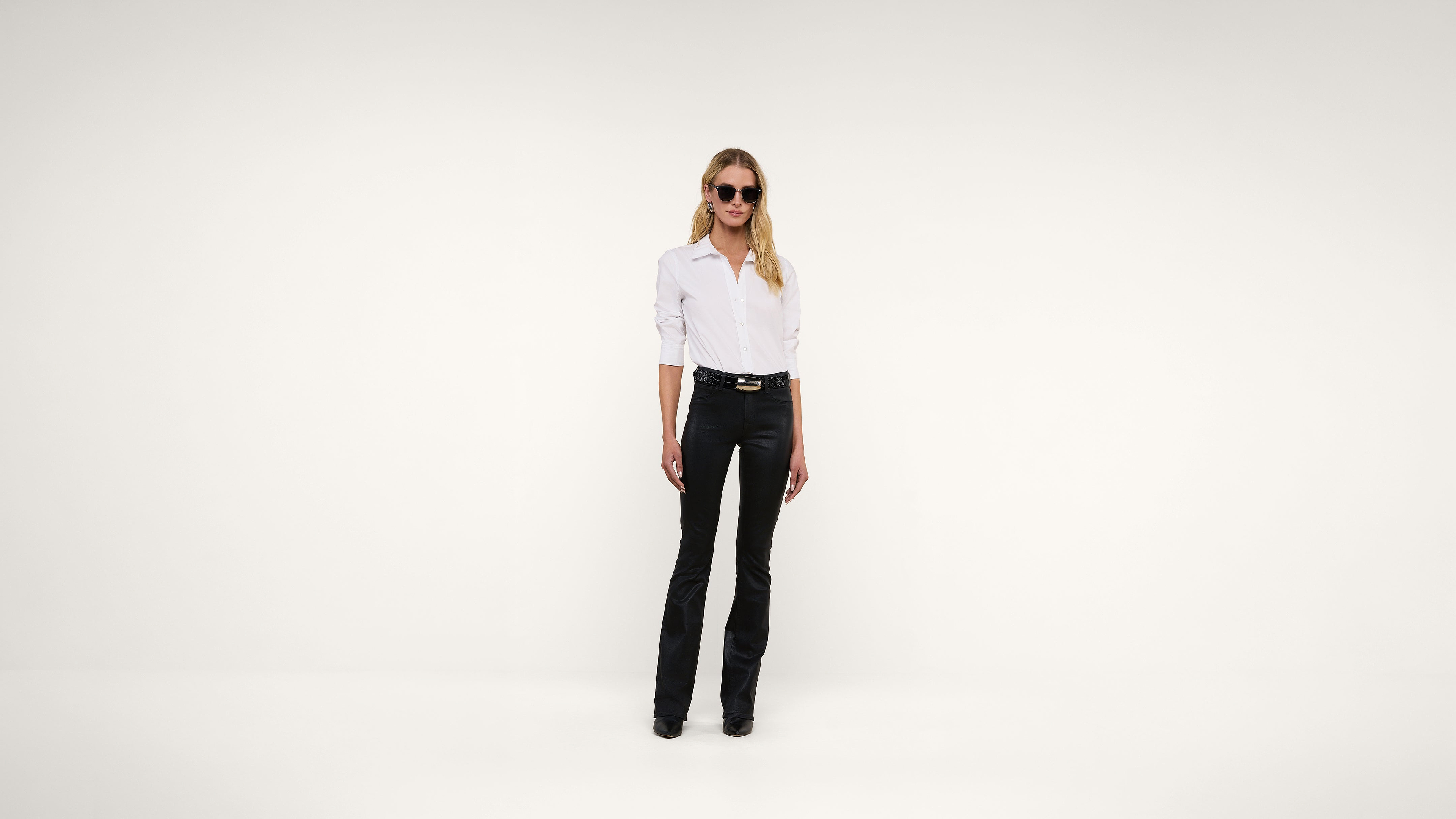 A woman wearing sunglasses, a white button-up shirt, and black pants stands against a plain white background.