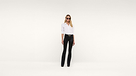 A woman wearing sunglasses, a white shirt, and black pants stands confidently against a plain white background, facing forward with her hands relaxed by her sides.